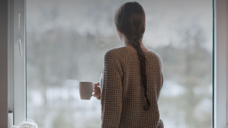Eine Frau sitzt eine Tasse Kaffee trinkend vor dem Fenster und schaut raus